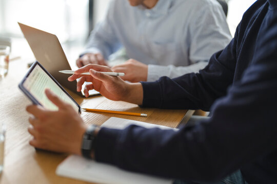 Two business colleagues collaborate in an office, reviewing documents on a digital tablet with a stylus while working together at a wooden table with an open laptop.