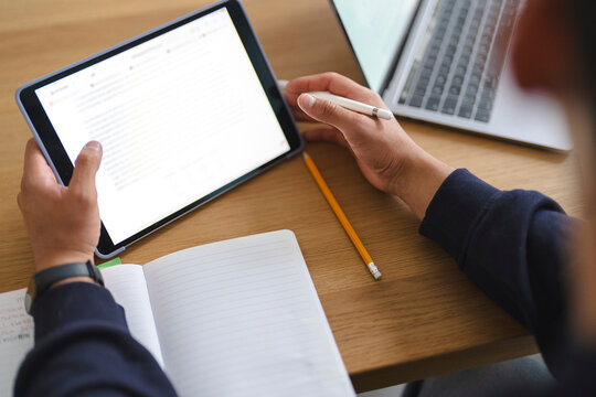 A person is working at a wooden desk, reading an online article on a digital tablet while holding a stylus. A laptop and open notebook sit nearby during a study session.