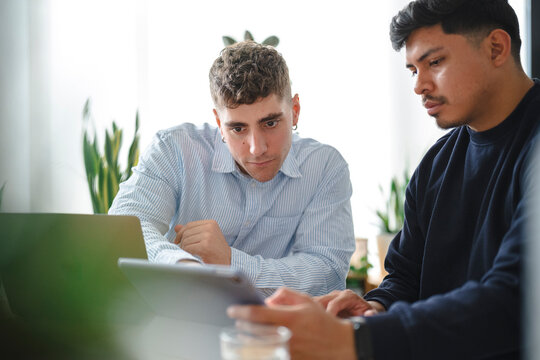 Two young male colleagues sit together at an office desk, collaborating on a project. They are looking closely at a digital tablet screen while working with a laptop.