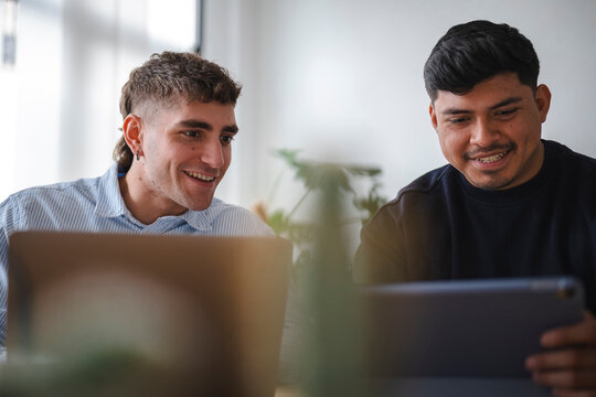 Two young male colleagues sit together at a wooden desk in a bright office. They smile while collaborating on a project using a laptop computer and a digital tablet.