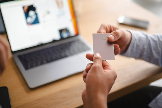 A man sitting at a wooden desk holds a credit card in his hands. He is shopping online using a laptop computer placed in front of him in a brightly lit room.