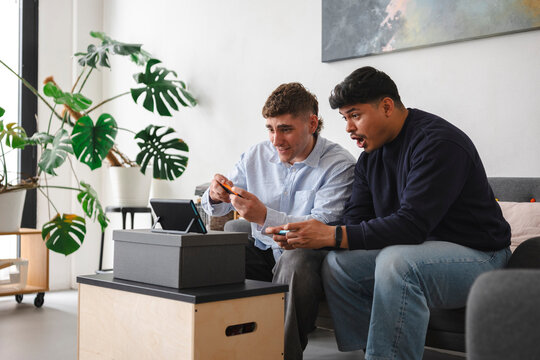 Two young male friends sitting together on a living room sofa, playing a video game console setup and reacting with excitement and surprise during an intense match.