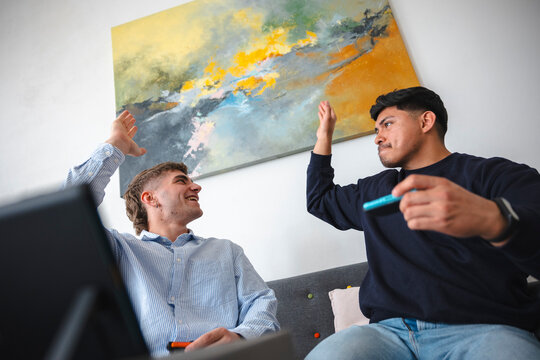 Two young men are sitting on a couch in a modern office or living room, giving each other a joyful high five while smiling. They are celebrating a successful project.