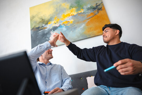 Two young male friends are sitting together on a grey couch in a bright living room, smiling and giving each other a high five while playing video games.