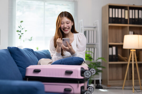 Happy woman packing luggage for travel planning vacation. Joyful Asian woman preparing for a trip, sitting on a sofa with a pink suitcase, celebrating travel bookings on smartphone.