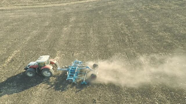 Sideways aerial drone flight tracking parallel to a tractor cultivating agricultural soil and kicking up a large dust cloud outdoors on a beautiful bright sunny summer day in countryside