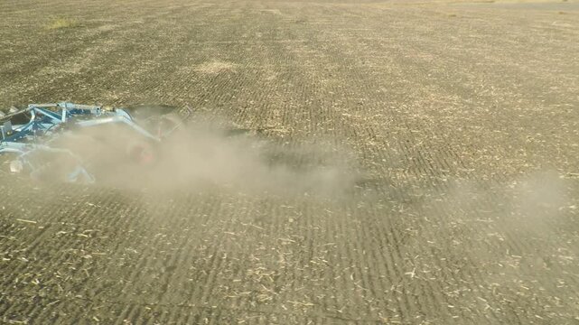 Sideways low angle drone flight tracking parallel to a tractor harrowing a finely plowed field, aerial view of farming machinery tilling soil and kicking up a large dust cloud on a sunny day