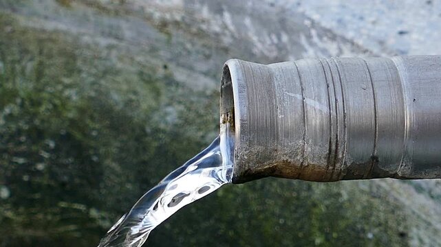 Macro profile shot of a roadside fountain stainless steel spout with flowing fresh water, side view of clean drinking water pouring down into a stone trough basin, video with audio included