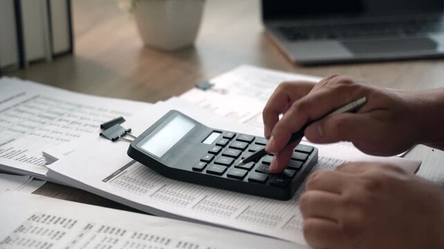 Close-up professional using calculator and pen to review financial documents, spreadsheets and business reports, checking figures, expenses and totals for accurate financial review.