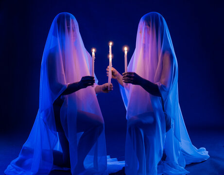 Two veiled kneeling figures hold candles in blue-lit dark studio