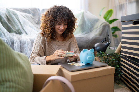 White woman counting money amidst packing boxes and piggy bank