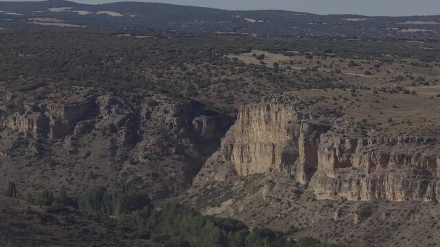 An aerial view shows layered cliffs above a tree-lined valley, with scrub-covered plateaus and distant fields in Aragon, Spain.