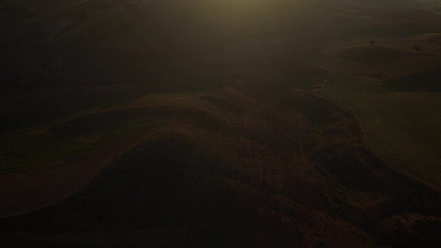 An aerial view reveals cultivated hills, curved crop rows and scattered trees beneath a low sun, with haze along the distant horizon in Aragon, Spain.