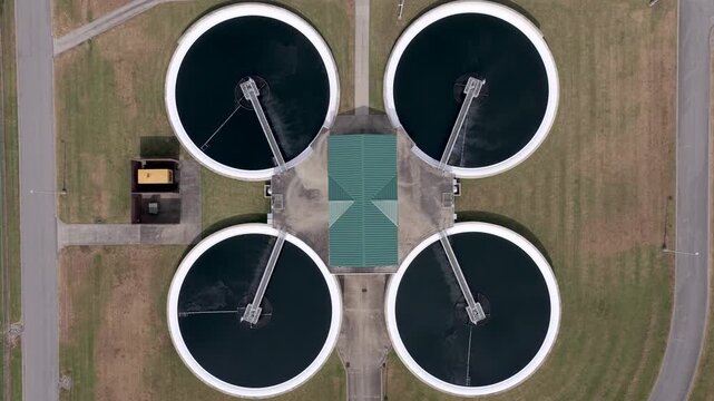 Four round clarifier basins with dark water and rotating skimmer bridges surround a green roofed central building at a wastewater treatment plant, lawns and paved paths framing them. Aerial top down