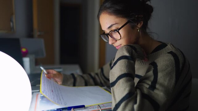 Focused teenage girl with glasses studying at her desk late at night. A diligent student concentrating on her notes for an exam preparation