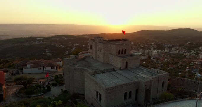 An elevated moving view passes Skanderbeg Museum at sunset in Kruje, Albania. An Albanian flag flies above the building, with red rooftops, hills and a hazy plain beyond.