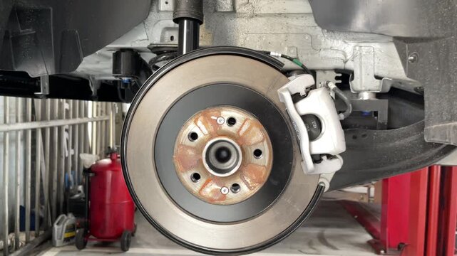 A widening view shows a car's exposed brake rotor, caliper and wheel hub, with suspension components inside the wheel well and workshop equipment behind.