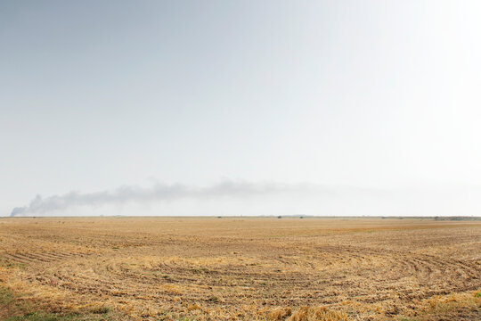Wide Dry Harvest Field Under Pale Sky With Distant Smoke Plume on the Horizon