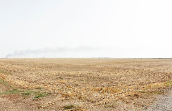 Dry Harvested Field Under Pale Sky With Distant Smoke On Flat Rural Farmland