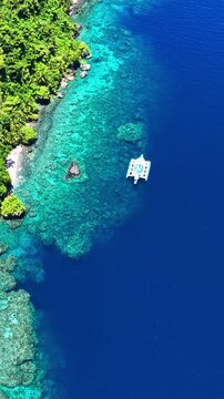 Aerial view of a white catamaran boat floating in the turquoise waters of Port Barton Beach, with lush green tropical vegetation along the coastline and deep blue sea, Mimaropa, Philippines.