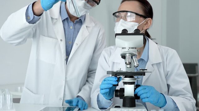 Two biotechnology professionals examining biological samples with a microscope and using a digital tablet for data entry during a scientific experiment in a life science lab