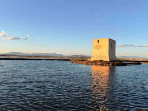 Quadratischer Steinturm auf Schilfinsel in ruhiger Lagune; goldenes Licht wirft warme Reflexion ins Wasser. Weit im Hintergrund sanfte Bergkette unter klarem, fast wolkenlosem Himmel.