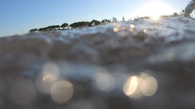 Half-underwater view of waves at the shoreline with sun glare, palm trees and closed beach parasols in the background, French Riviera, France.