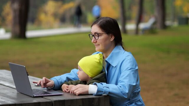 Busy Mother Balances Remote Work Using Laptop Computer While Holding Toddler Son In Autumn Park, Facing Parental Burnout And Exhaustion During Hectic Multitasking Outdoor Routine In Daytime