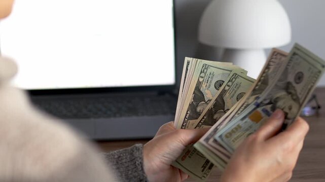 Woman hands count a stack of US dollar bills at a desk with an open laptop in the background. Remote work income concept about freelance earnings, online business profit and salary paid in cash.