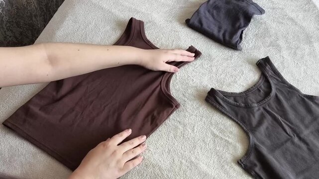 Woman neatly folding T-shirts and tank tops after laundry, illustrating clothing care, household organization, and preparation of summer garments for storage in a wardrobe.