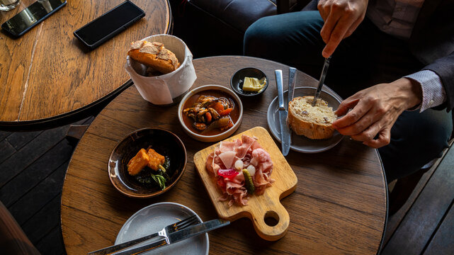 Man spreading butter on sourdough bread at wooden table with prosciutto ham, stew, and tapas in restaurant
