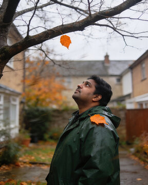 A man in a green raincoat gazing up at the last hanging leaf, reflecting the nostalgic connection between human and autumn weather.