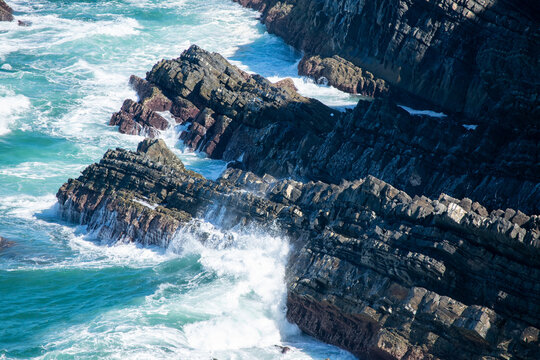 Powerful Turquoise Waves Crashing Against Dark Stratified Coastal Cliffs