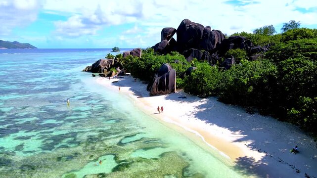 Bright daytime view of a tropical beach on La Digue Island, Seychelles, with visitors walking beside clear turquoise water, pale sand and dramatic granite boulders.