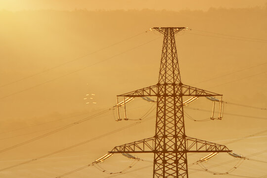 Close up of high voltage power transmission pylon top section with glass insulators and wires in thick golden fog. Industrial electric serves