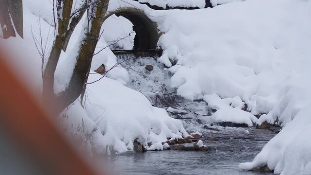 Water flows from culvert under snowy bridge in winter park. Moving water contrast with static snow creates dynamic natural composition