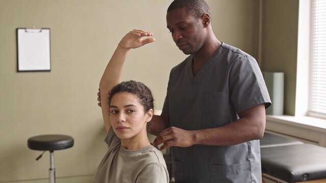 Assessing clinician in scrubs supporting patient raising arm in therapy room, with table