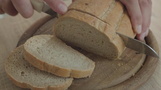 High angle view of male hands slicing crusty artisanal bread loaf on wooden cutting board, slow motion food preparation in kitchen