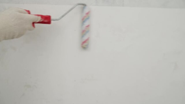 A close up of a hand in a glove using a paint roller to apply adhesive or primer to a prepared wall. Home improvement, DIY repair, and interior finishing concept. Construction work in progress