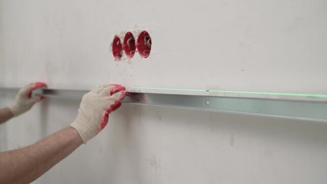 Close up of a construction worker in gloves mounting a galvanized metal stud. A green laser level line guides the precise horizontal installation on a plaster wall. Renovation and building concept