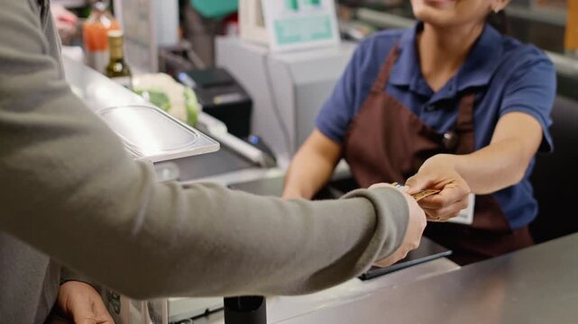 Unrecognizable customer paying with contactless credit card at checkout counter while female cashier handing discount coupons. Ideal for retail banking, electronic commerce, supermarket advertising