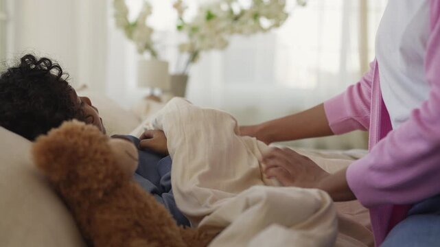 African American boy falls asleep in bed as caring mother adjusts blanket