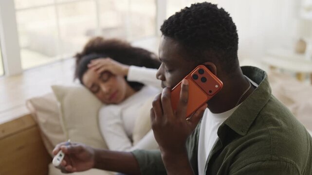 Sick woman holds forehead as husband discusses symptoms with doctor by phone