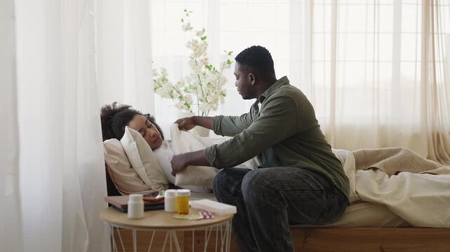 Compassionate Black husband cares for sick wife resting in bed, recovery