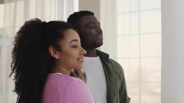 Smiling young Black couple hug while looking out window, planning future