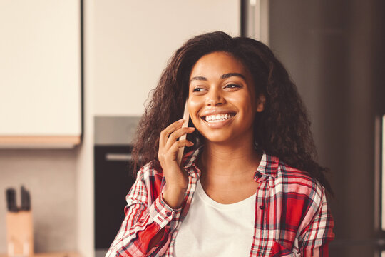 Happy Young Black Woman Smiling While Talking On Phone In Modern Kitchen, Engaged In Conversation. Enjoying Time In Home Setting, Expressing Positive Emotions And Connection. Communication Concept.