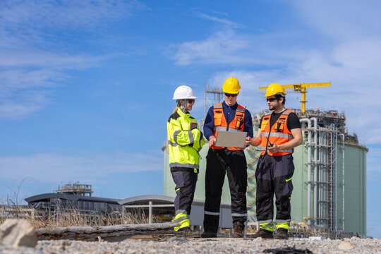 Group of industrial engineers using laptop computer while reviewing data near gas storage tank facility, Engineering team holding laptop and walkie talkie during site inspection at refinery plant