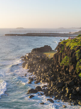 Aerial view of Fingal Head featuring rugged basalt rock formations, crashing ocean waves, and a distant jetty under soft golden hour light in Fingal Head, New South Wales, Australia.