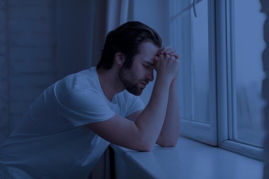 Worried Young Caucasian Man With Hands Clasped Facing Window In Indoor Setting, Expressing Stress And Anxiety, Reflecting On Life While Looking Outside In A Calm Yet Pensive Moment. Mental Wellness