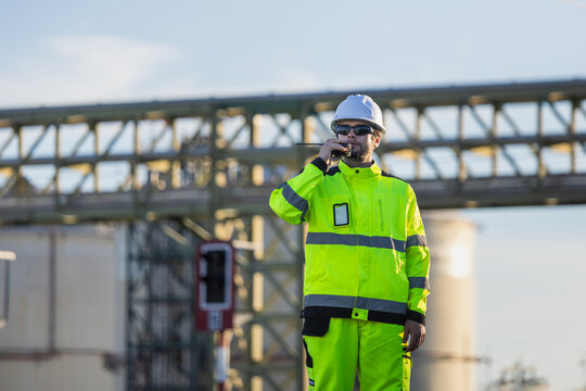 Engineer man using walkie talkie for communication at refinery plant, Industrial worker in high-visibility safety jacket talking on walkie talkie radio during site inspection outdoors